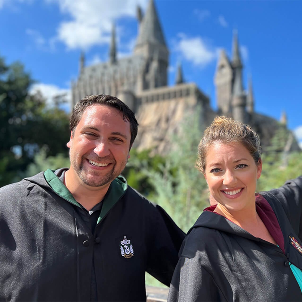 The hosts of the Discover Universal Podcast in Slytherin™ and Gryffindor™ robes posing in front of Hogwarts™ castle at Universal Islands of Adventure.