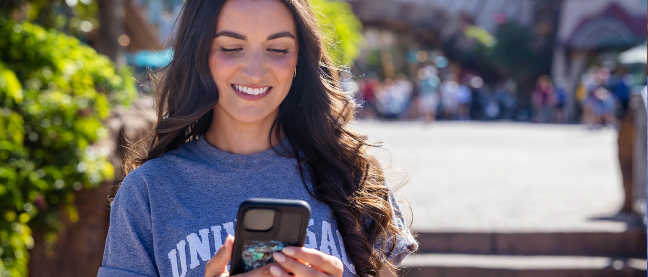 A Guest in a blue Universal T-shirt smiling while using a mobile phone with sunny open theme park space behind her.