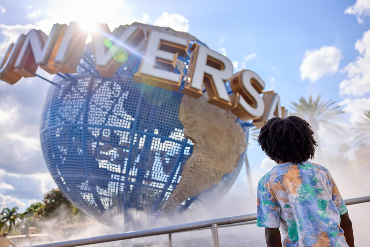 A young Guest viewed from behind gazing up at the iconic Universal globe bathed in sunlight and water mist.