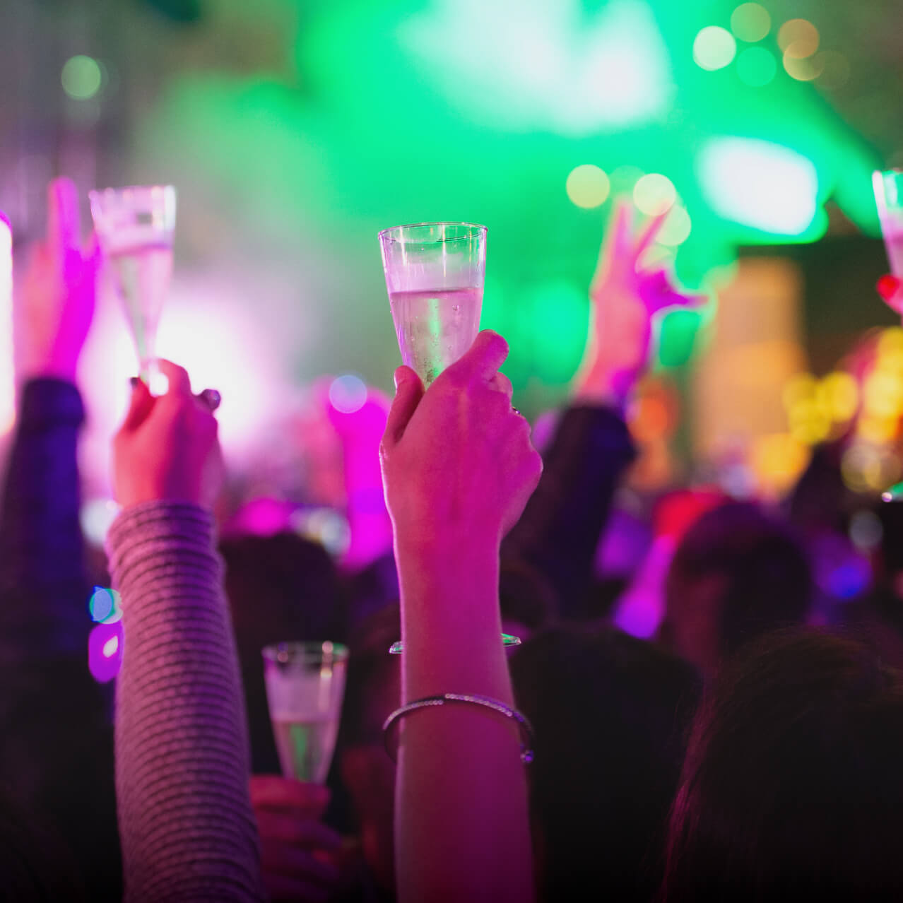 Multiple hands raising their glasses during a champagne toast on New Years Eve with out-of-focus lights in the background.