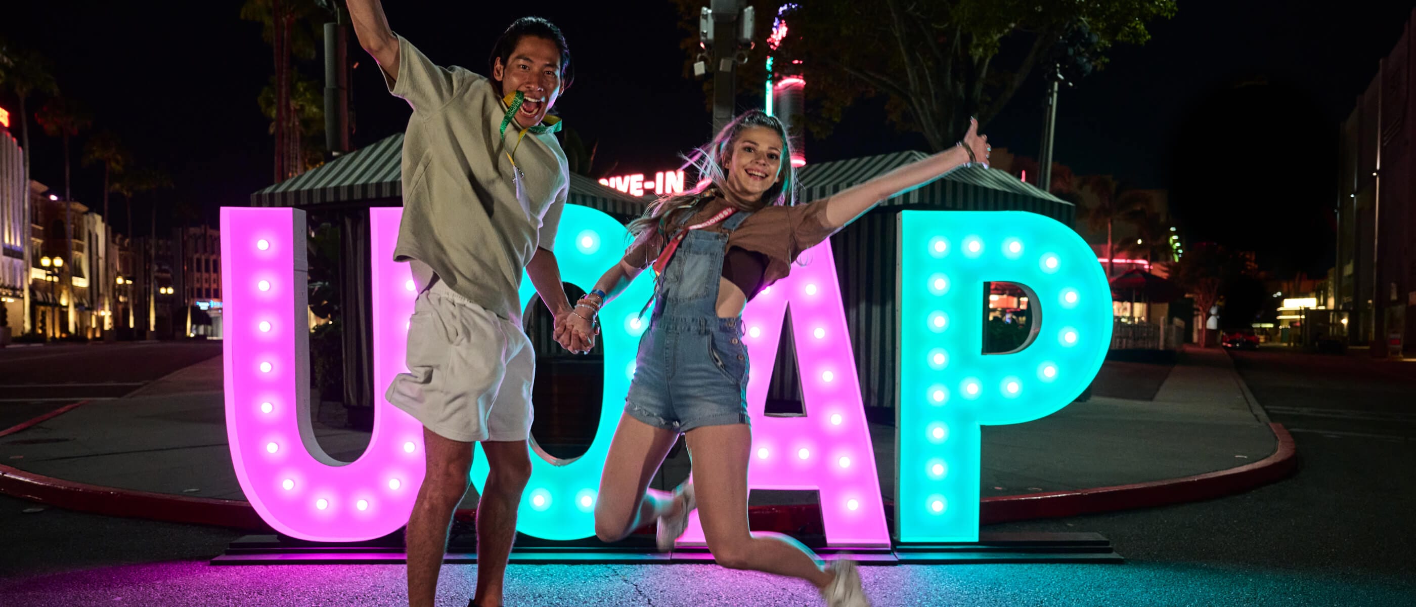 Two excited lanyard-wearing Passholders jumping in the air while holding hands in front of a large “UOAP” sign in pink and green lights.