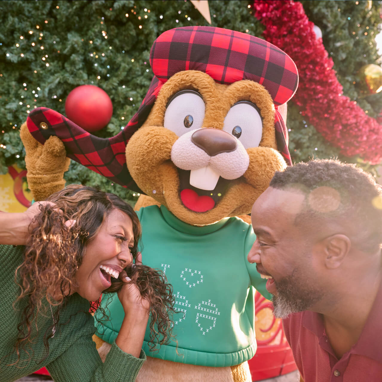 A couple smiling and laughing as they pose with Earl the Squirrel in front of festive holiday decorations.