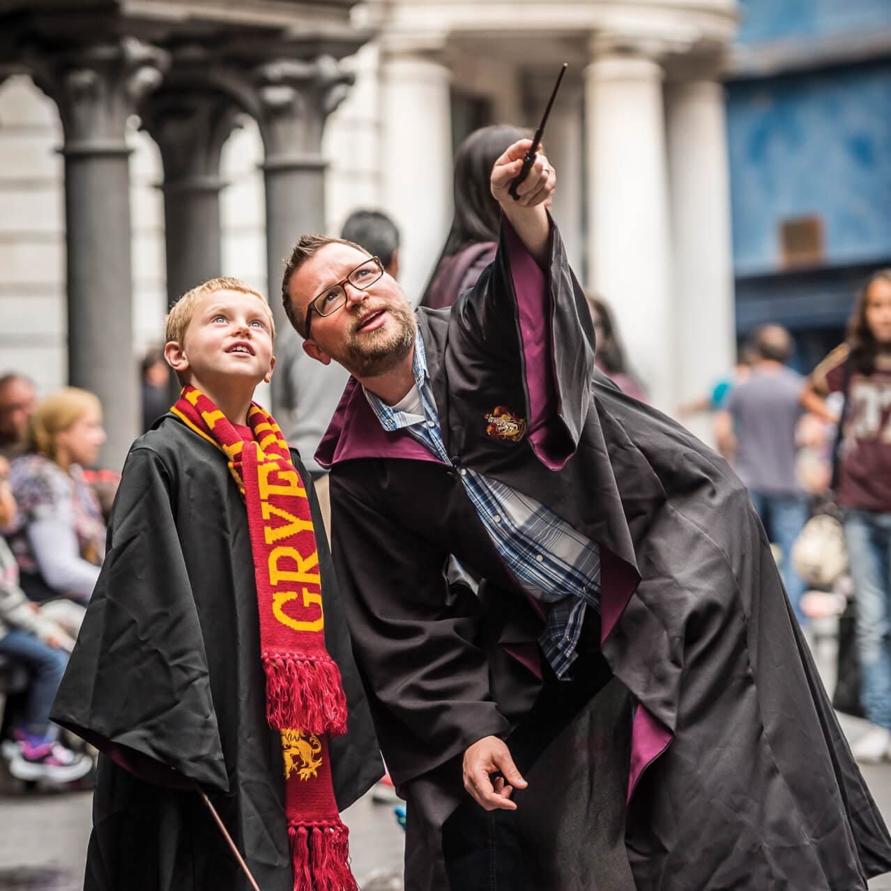 A father and child in wizarding robes gaze up to where the father’s wand is pointing, with Gringotts Bank in the background.