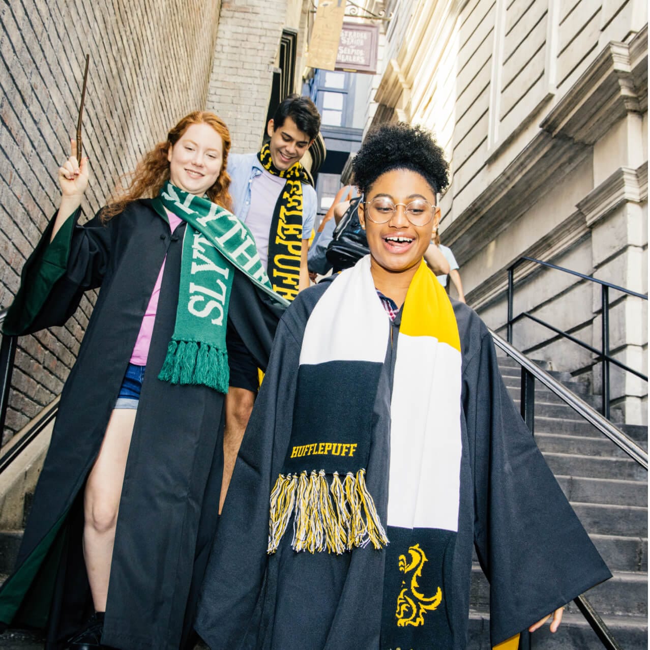 A woman wearing a Hogwarts cloak and a Slytherin scarf holds a wand as she and her two friends walk down a set of stairs.