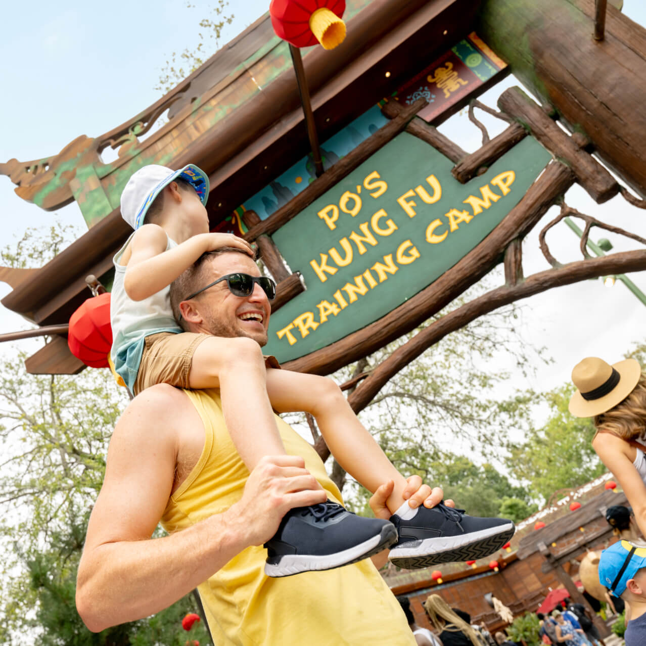 A&nbsp;smiling&nbsp;father&nbsp;with&nbsp;a&nbsp;child&nbsp;on&nbsp;his&nbsp;shoulders&nbsp;walks&nbsp;past&nbsp;the&nbsp;wooden&nbsp;entrance&nbsp;sign&nbsp;for&nbsp;Po’s&nbsp;Kung&nbsp;Fu&nbsp;Training&nbsp;Camp.