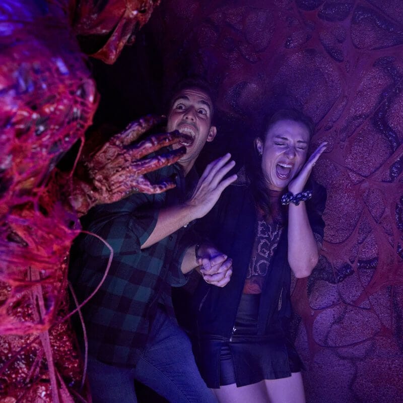 A couple holding hands and screaming in terror as a slimy creature reaches its decaying hand out toward them in the darkness.