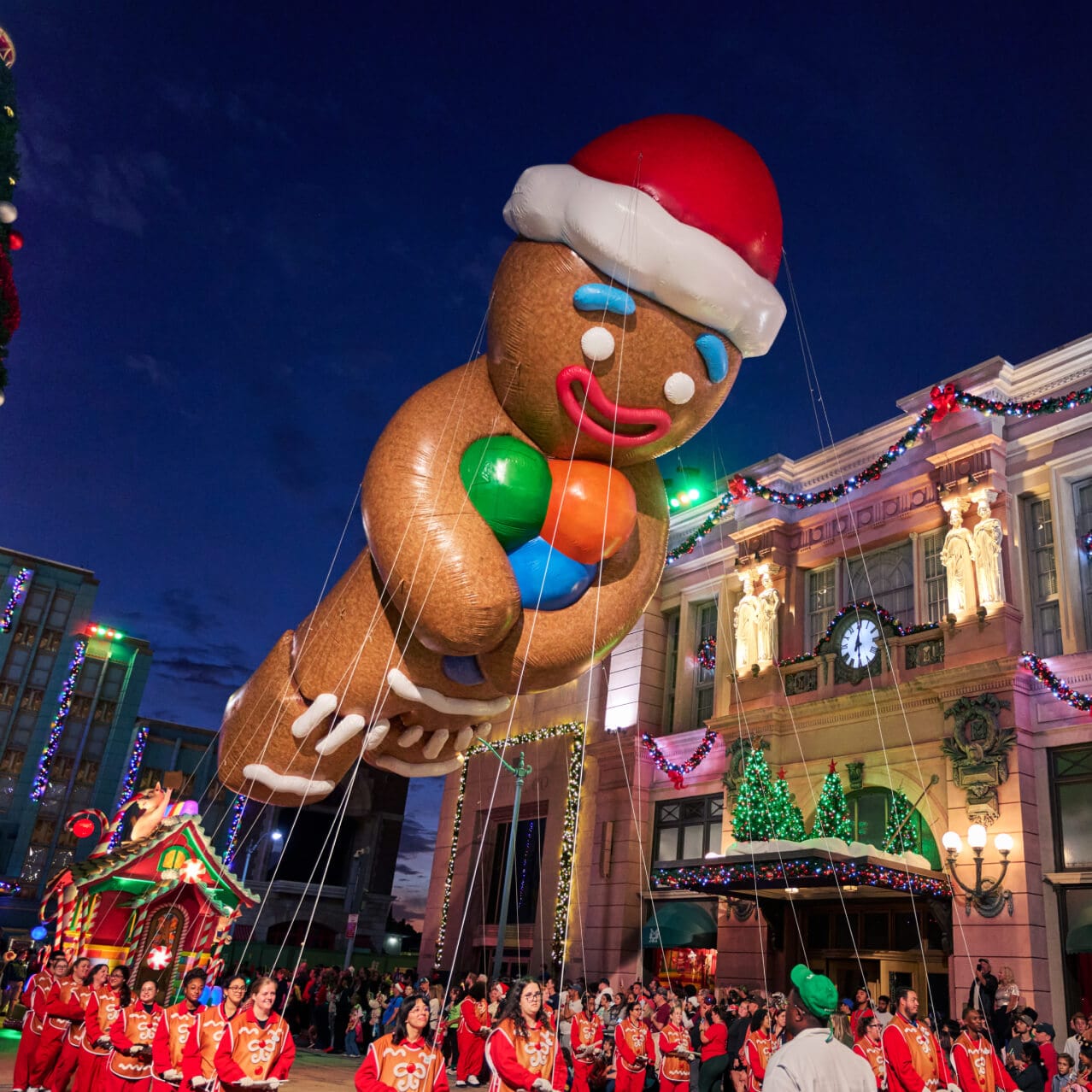 A large float balloon of the Gingerbread Man in a Santa hat holding colorful candies passing festively decorated buildings. &nbsp;