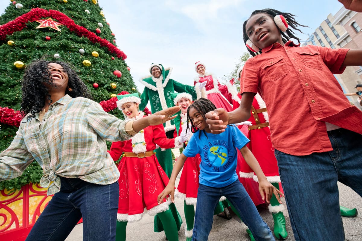 A mom and kids dance with some elves dressed in festive reds and greens alongside a large Christmas tree at Universal Studios Florida.