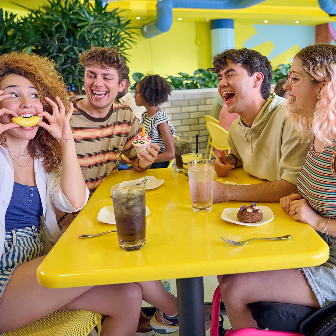 A group of friends, one using a wheelchair, laughing over snacks at a table at Illumination’s Minion Café.