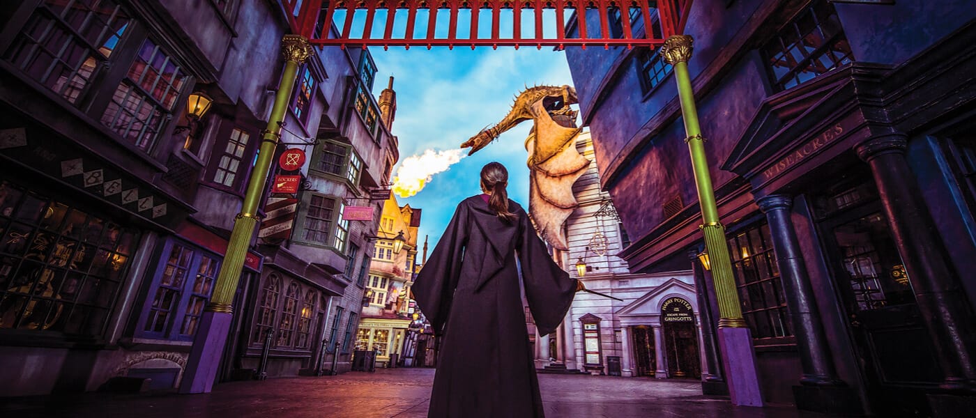 A girl in a wizard robe stands alone in The Wizarding World of Harry Potter™ - Diagon Alley™, as the dragon blows fire.