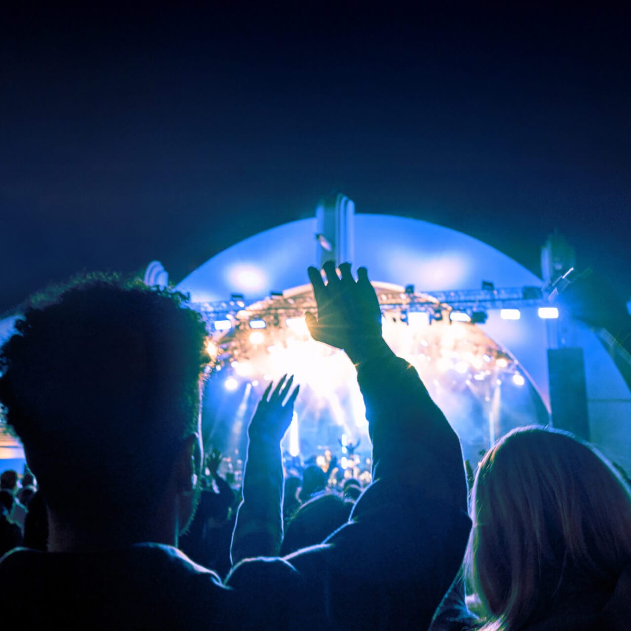 A brightly illuminated outdoor stage during a nighttime concert; in the foreground silhouetted concert goers wave their arms.