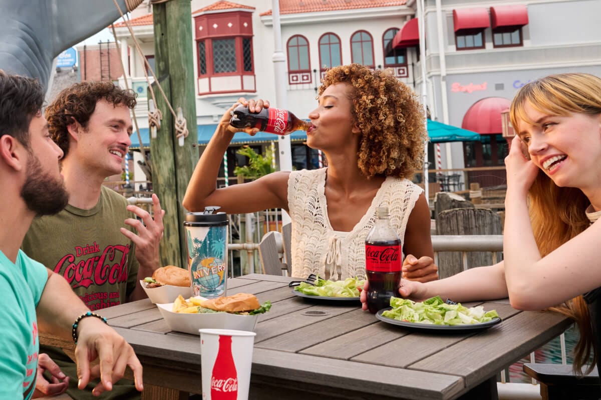 Happy friends enjoying food and Coke beverages at an outdoor table by the Jaws shark prop and Fisherman’s Wharf buildings.