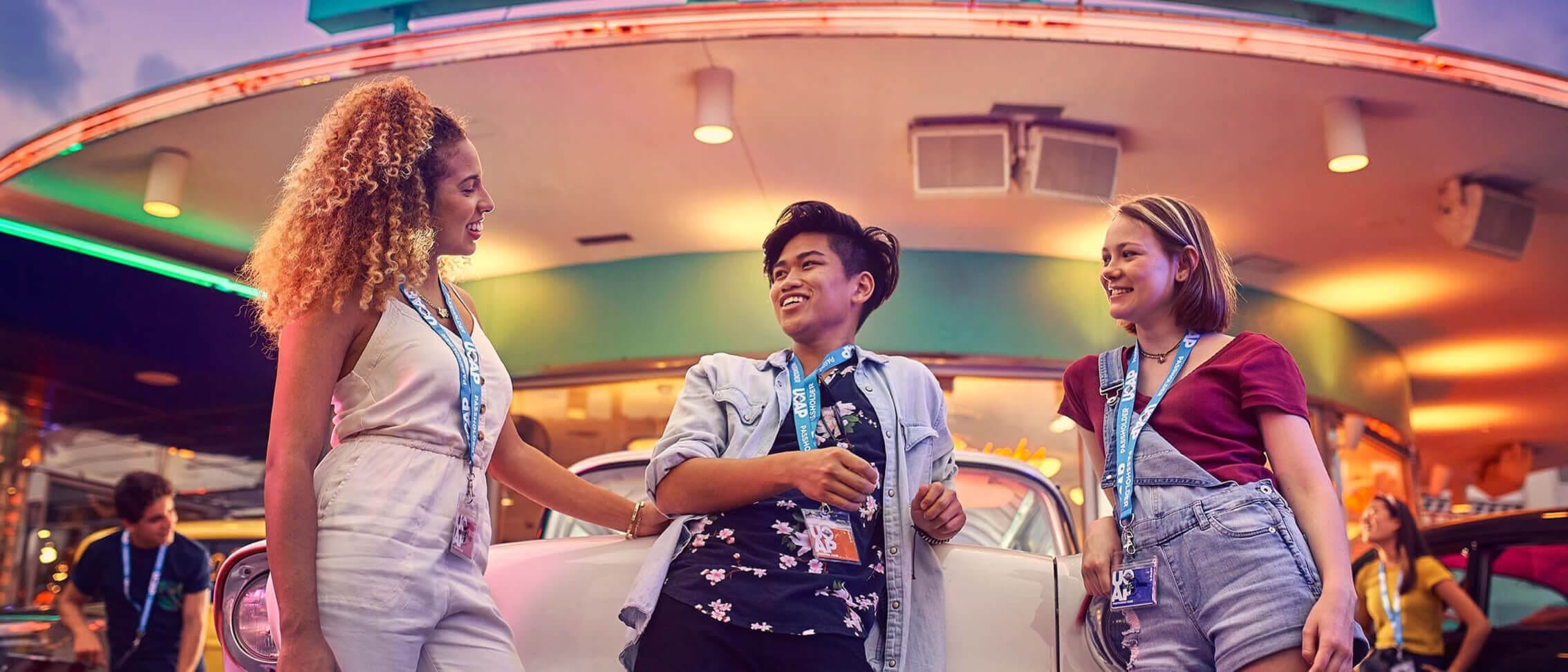 Three lanyard-wearing Passholders smiling while leaning on a classic car outside Mel’s Drive-In at Universal Studios Florida.