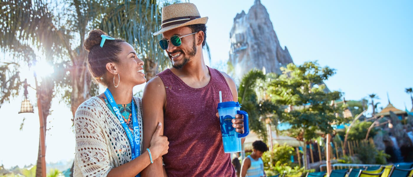 A couple smiles at each other in front of the volcano at Universal's Volcano Bay. The woman is wearing a UOAP lanyard and the man is holding a UOAP cup.