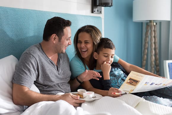 A happy family greets the morning as they sit up in their hotel bed. Dad has a breakfast plate on his lap, while a laughing Mom and their young son look over a brochure.