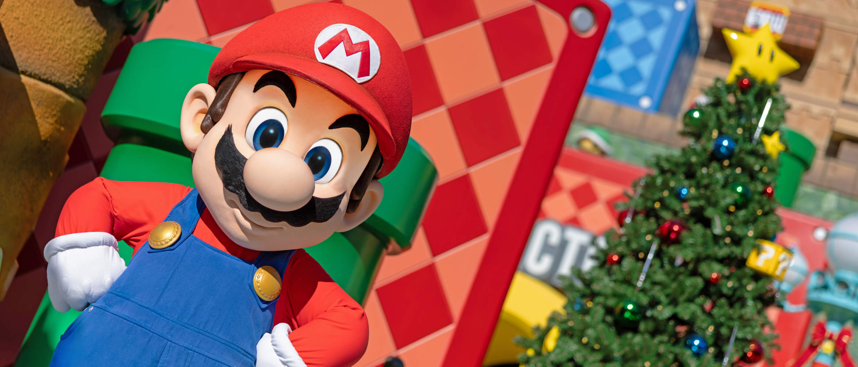 Mario with his hands on his hips in front of a green pipe and a large Christmas tree in the colorful SUPER NINTENDO WORLD
