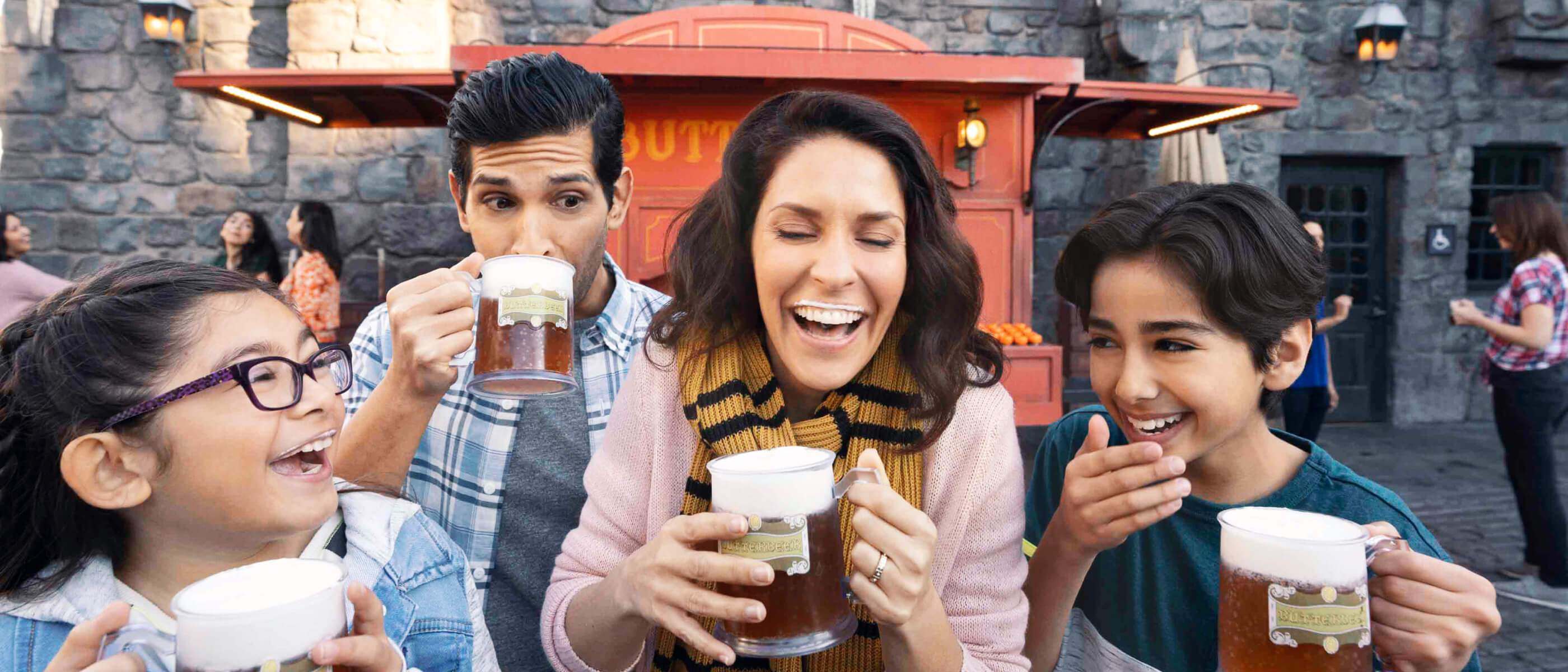 A family laughing with foam mustaches as they enjoy Butterbeer in front of a red cart on the streets of the Wizarding World.
