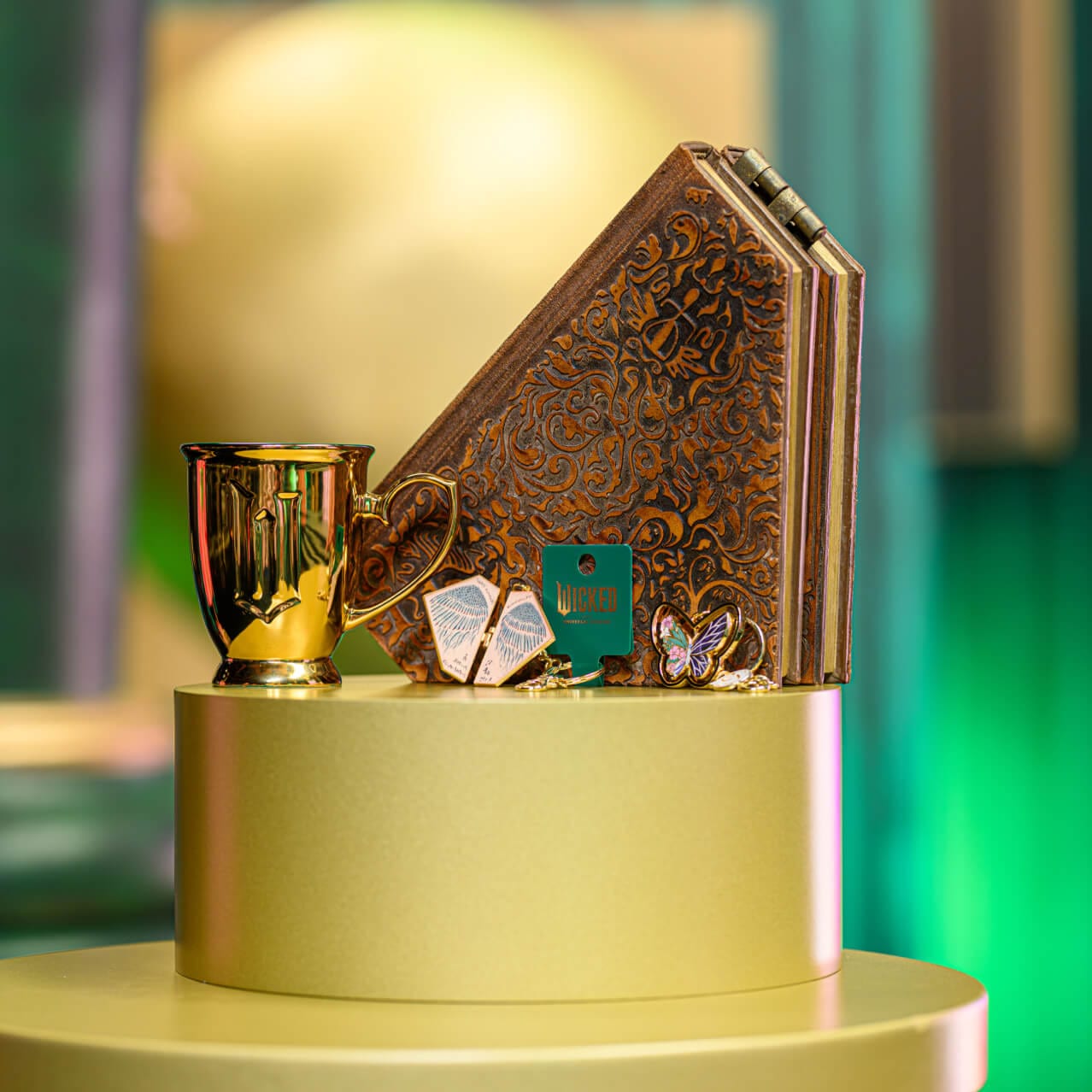 Wicked: For Good merchandise including a gold mug, a butterfly-shaped keychain and a keychain with wings.
