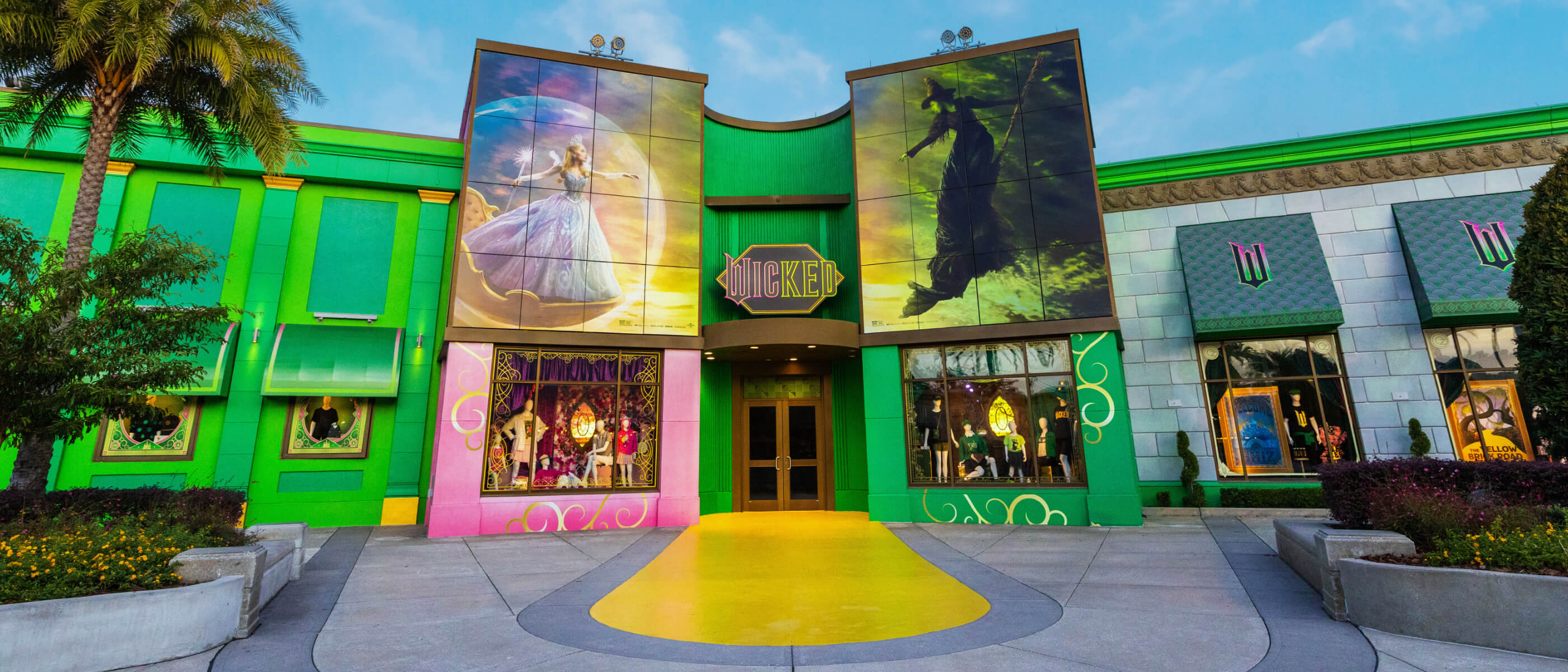 The front exterior of Wicked: The Experience with a yellow brick road entry and pink and green Glinda and Elphaba posters.