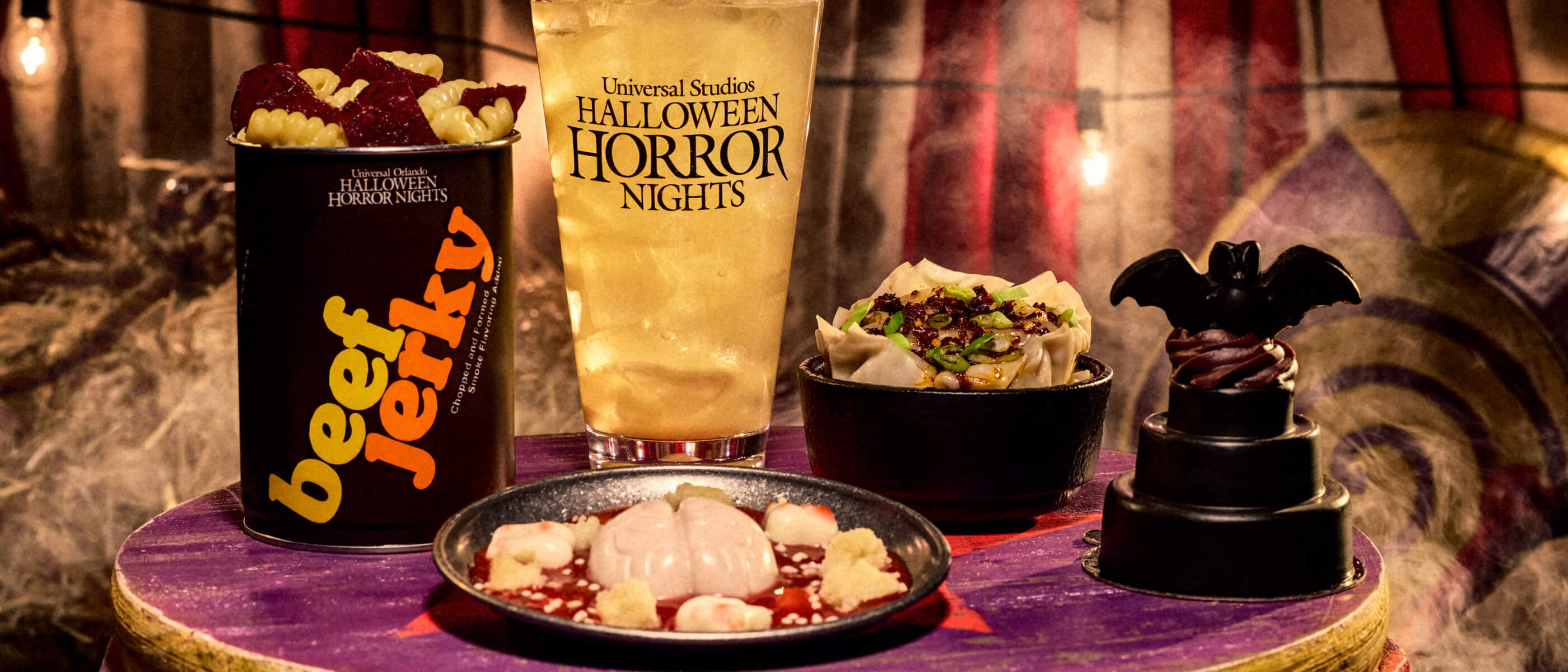 Assortment of Halloween Horror Nights food- creamy mac 'n' cheese, beef lasagna, custard, carrot cake and whiskey drink on a purple drum.