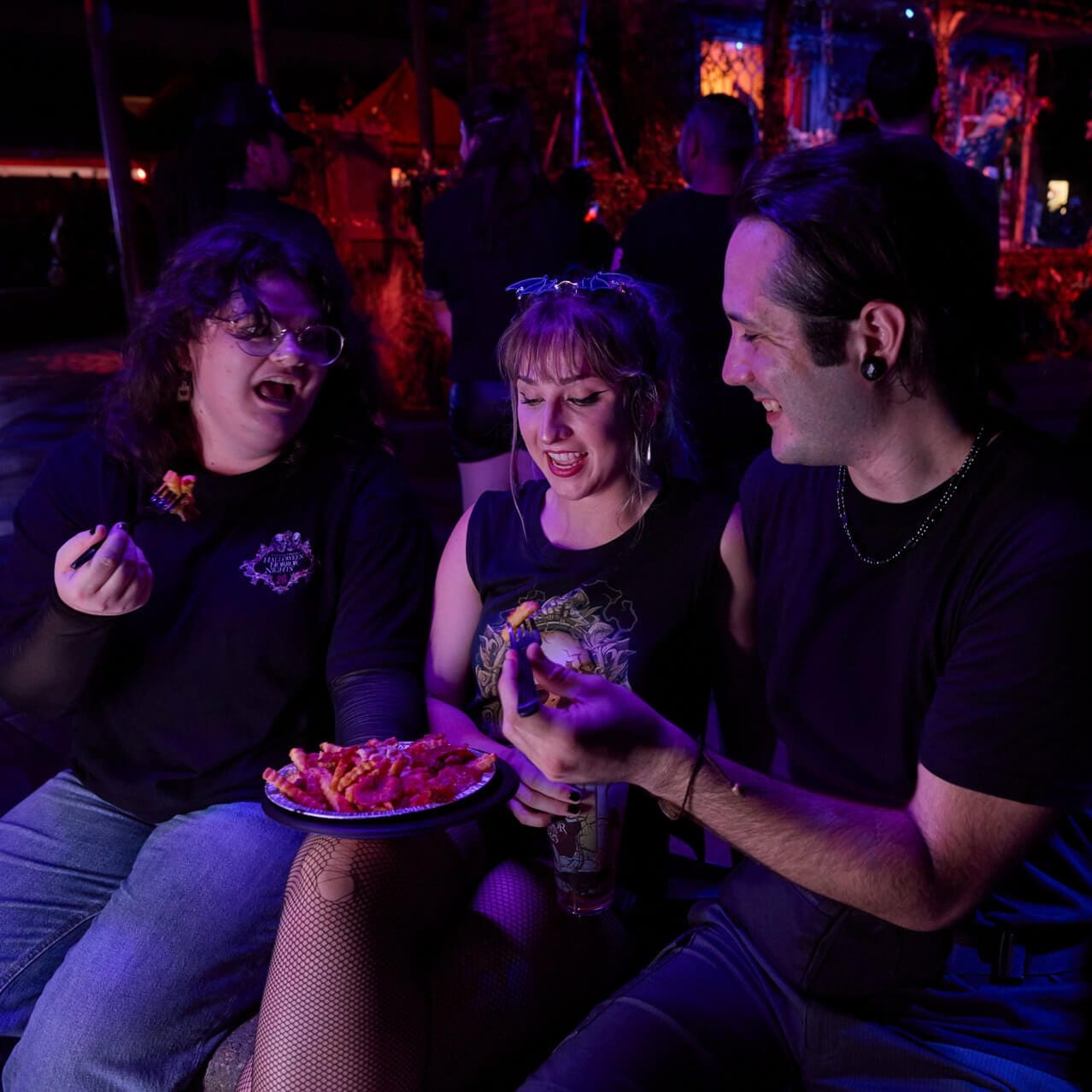 Smiling friends sitting together and sharing a plate of pizza fries with Halloween-themed lighting and décor behind them.