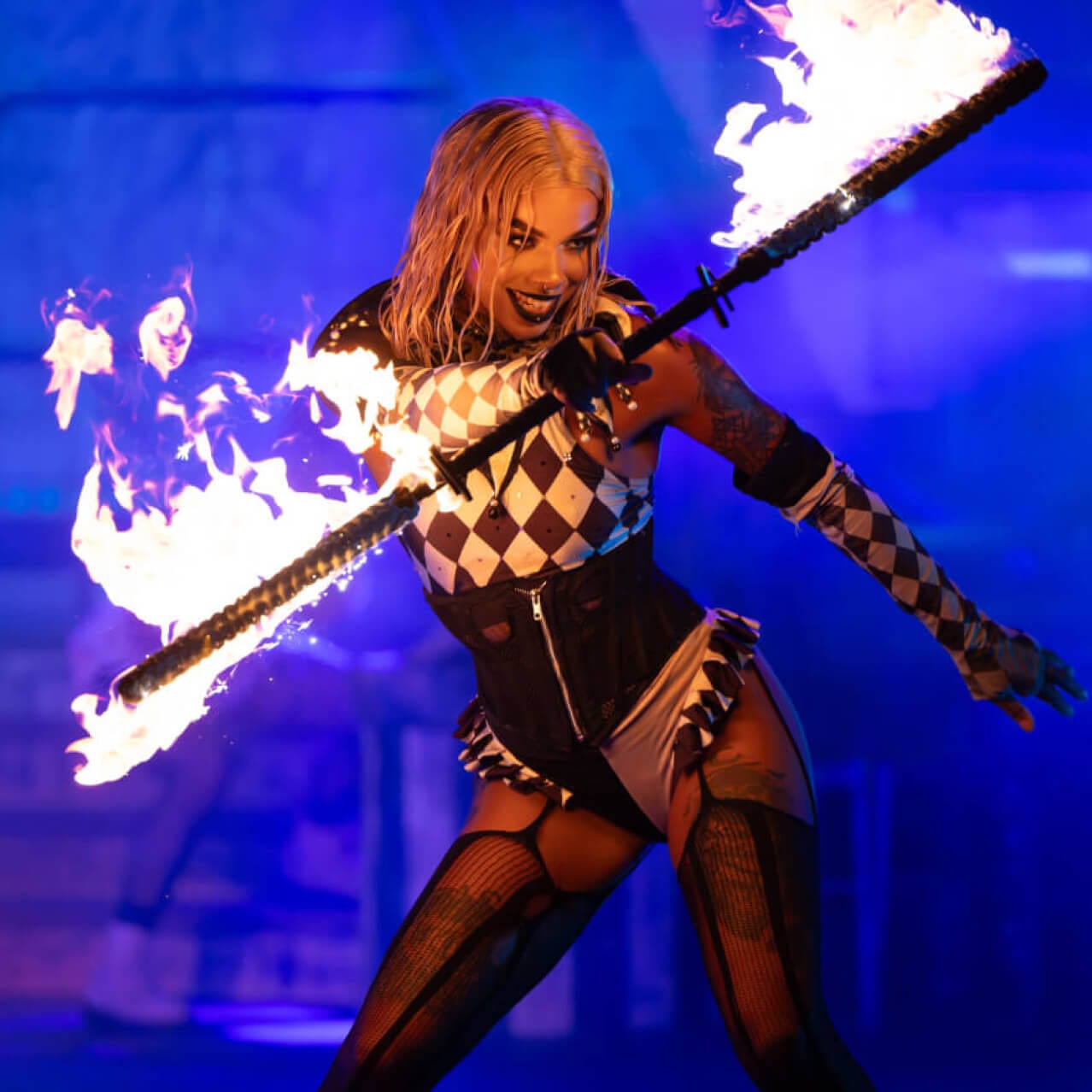 An onstage performer with black makeup and black-and-white checkered apparel twirling a long baton with flames at either end.