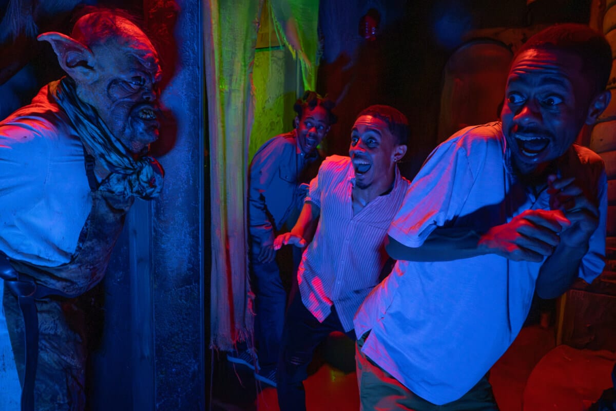 Friends recoiling in fear as a goblin-like scareactor with pointed ears and an apron emerges from the darkness.