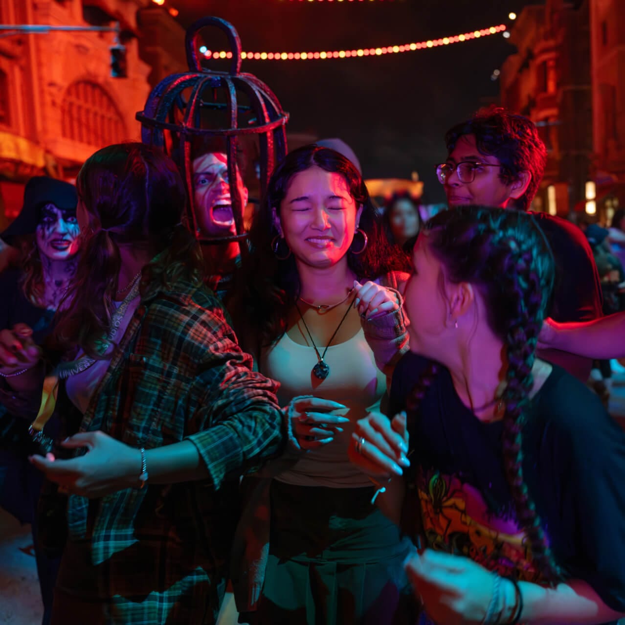 A group of anxious guests trying to avoid two menacing scareactors in the night streets, one with a medieval torture cage on its head.