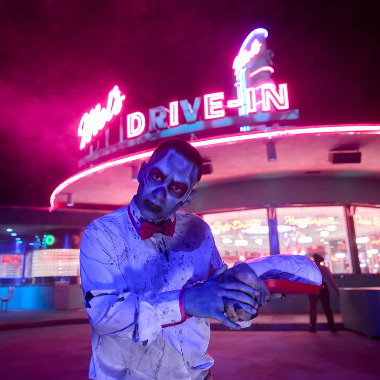 A zombie scareactor in tattered clothes standing outside Mel’s Drive-In under pink and blue neon lights.