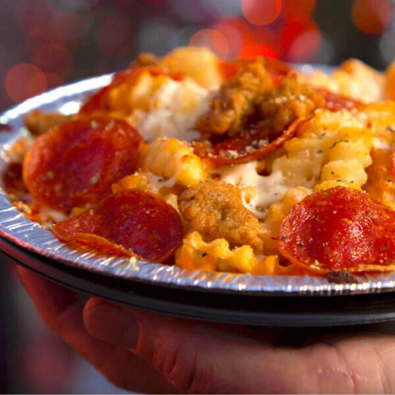 A hand holding a bowl of crinkle cut fries topped with marinara sauce, melted cheese, pepperoni and sausage.