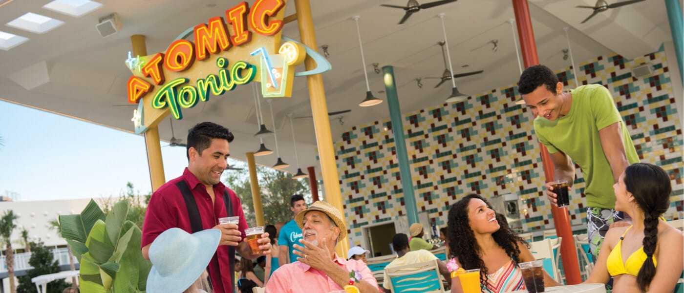 Un mesero le sirve a visitantes sentados en una mesa afuera de Atomic Tonic, un bar al aire libre estilo retro en Universal's Cabana Bay Beach Resort.