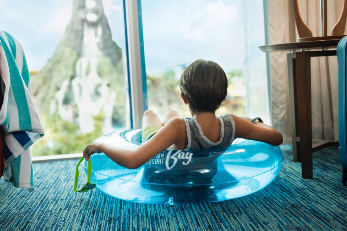 Un niño sentado con un salvavidas puesto en su habitación en Universal Cabana Bay Beach Resort, mientras disfruta de una vista de Universal Volcano Bay a través de la ventana.