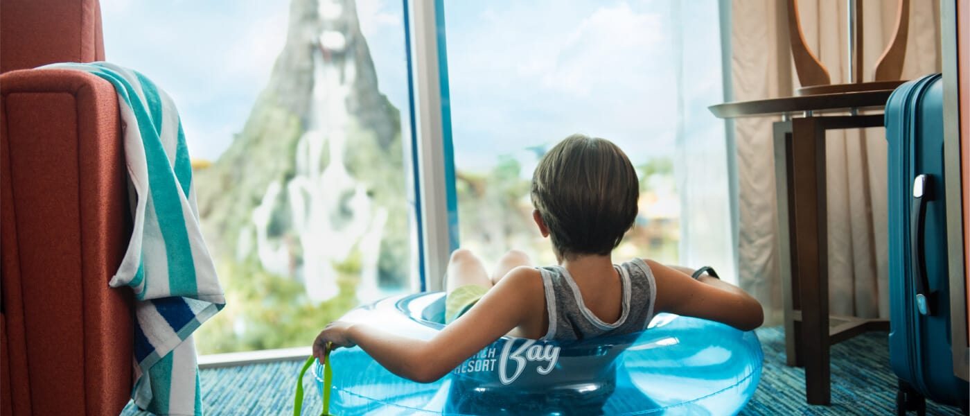 Un niño sentado con un salvavidas puesto en su habitación en Universal's Cabana Bay Beach Resort mientras disfruta de una vista de Universal's Volcano Bay a través de la ventana.