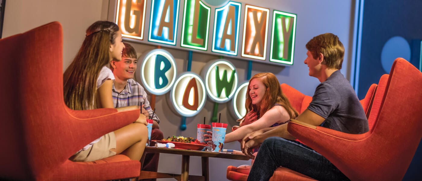 Una familia sentada en sillas estilo retro disfruta de una cena cerca del letrero iluminado de Galaxy Bowl en Universal's Cabana Bay Beach Resort.