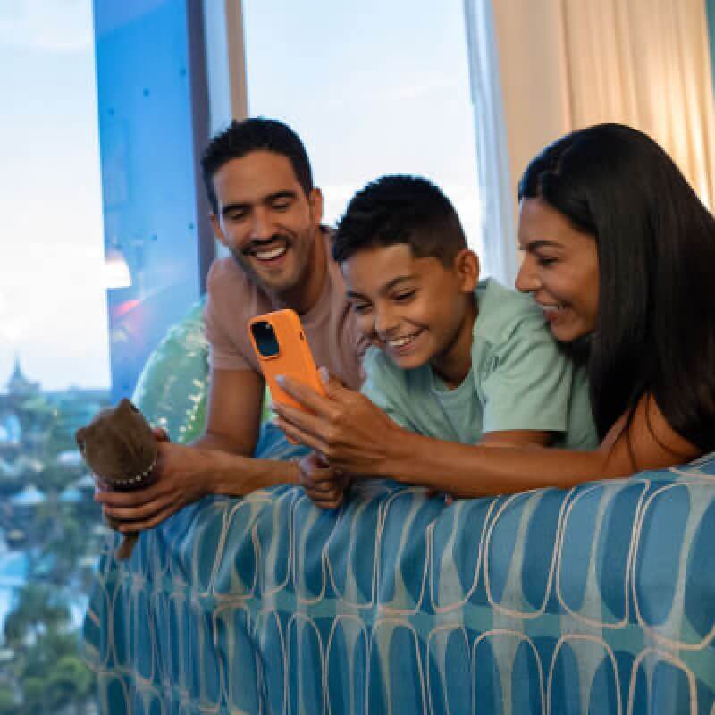 Una familia de tres ríe mirando un teléfono mientras descansan en una habitación de hotel en Universal's Cabana Bay Beach Resort.