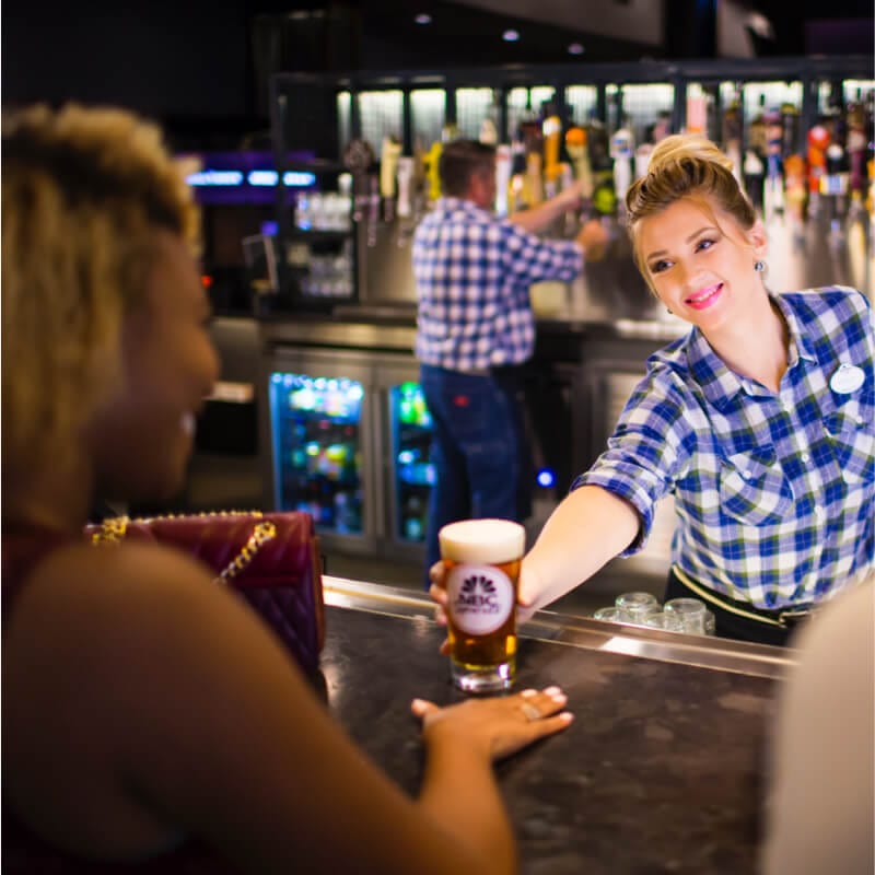 Un mesero vestido en una camisa azul con patrón de ajedrez le ofrece una cerveza a una mujer en NBC Sports Grill And Bar en Universal CityWalk Orlando.