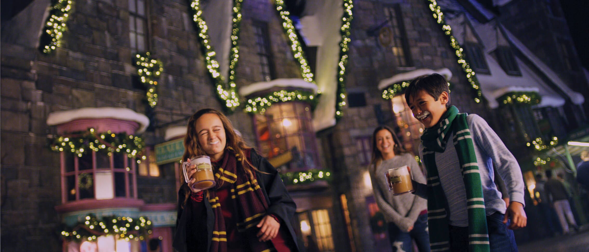 Una hermana, su hermano y su madre pasean en Navidad en The Wizarding World of Harry PotterTM - HogsmeadeTM mientras beben ButterbeerTM.