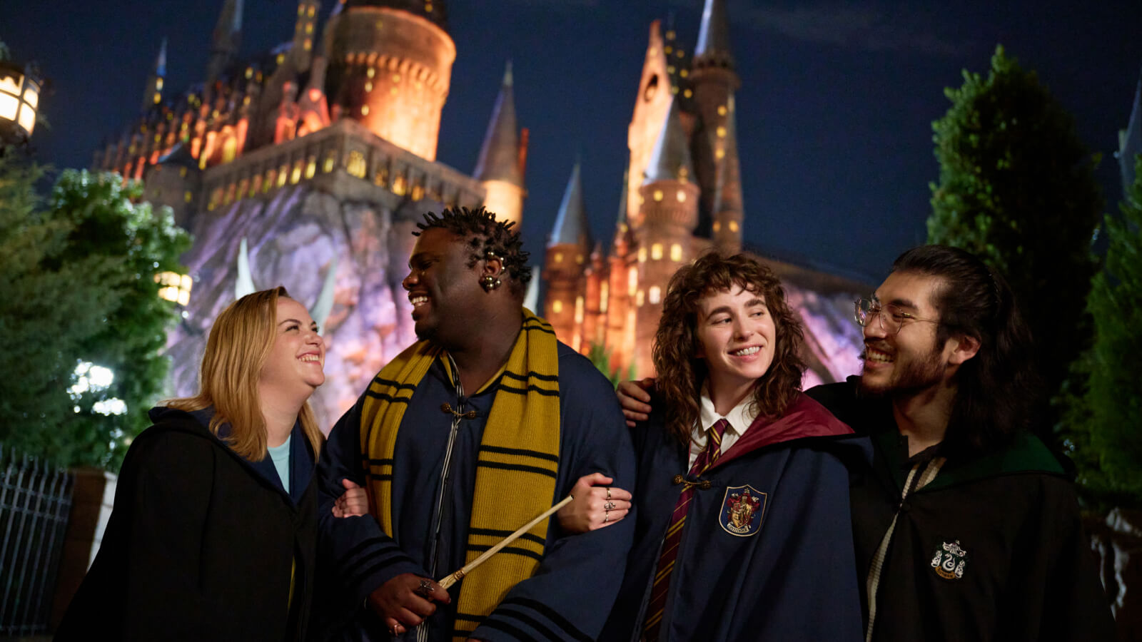 Cuatro amigos con túnicas y accesorios mágicos sonríen y se ríen frente al castillo de Hogwarts™ iluminado en la noche.