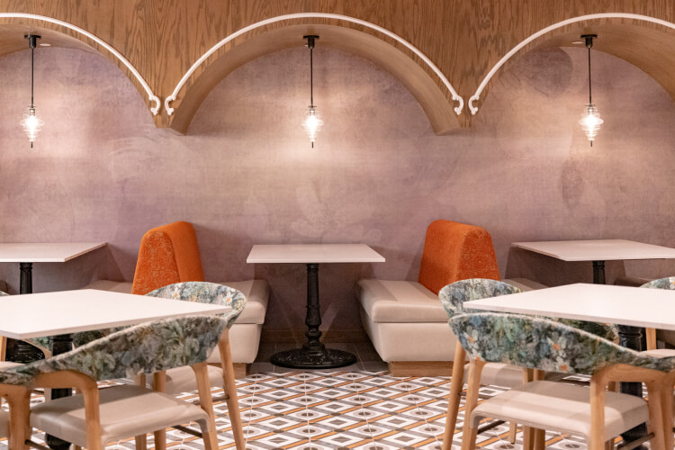 A warmly lit lounge area with recessed booths in the background and square tables with chairs in the foreground.