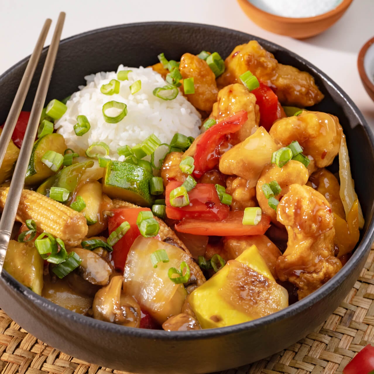 A bowl of crispy chicken, white rice, and vegetables on top of a bamboo placemat with metal chopsticks.