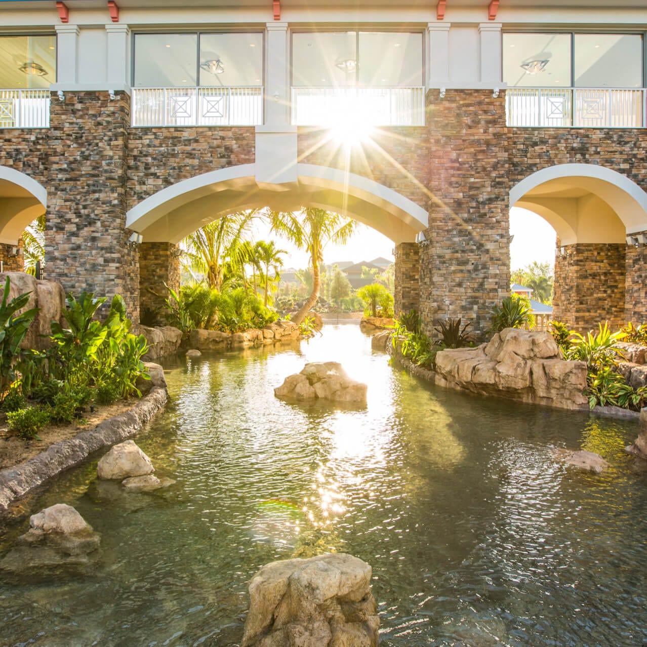 El sol brilla tenuemente a través de un puente cerrado que se extiende sobre un arroyo rodeado de palmeras, creando una pintoresca escena caribeña en el Loews Sapphire Falls Resort de Universal Orlando.