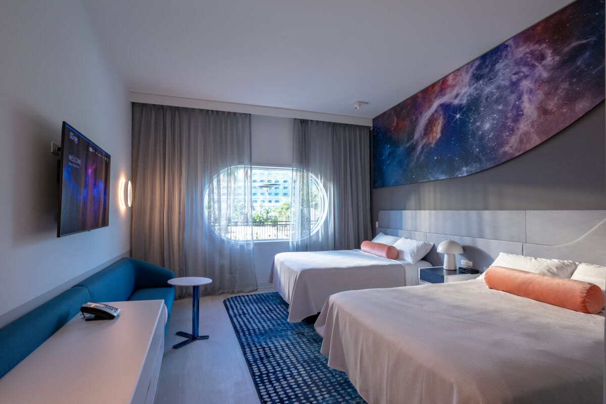 Habitación estándar de Universal Stella Nova Resort con dos camas queen y mesas de noche, sofá, mesa, vestidor, televisión y ventana ovalada en el fondo.