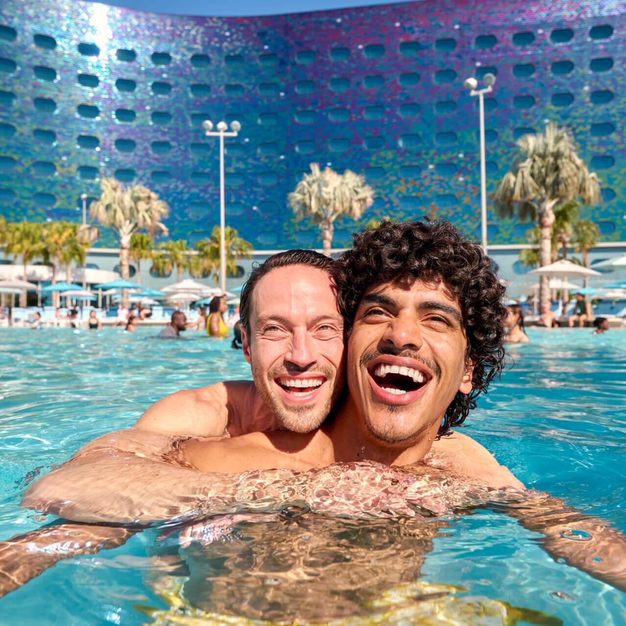 Una pareja riendo mientras disfrutan de un chapuzón en la piscina de Universal Terra Luna Resort, con la fachada de paneles del resort brillando en el fondo.