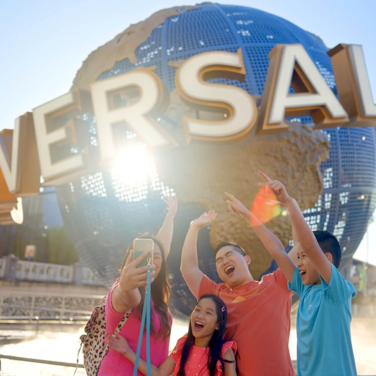 Una familia de cuatro integrantes levanta los brazos y sonríe para una selfi frente al icónico globo de Universal Orlando en Universal Orlando Resort.