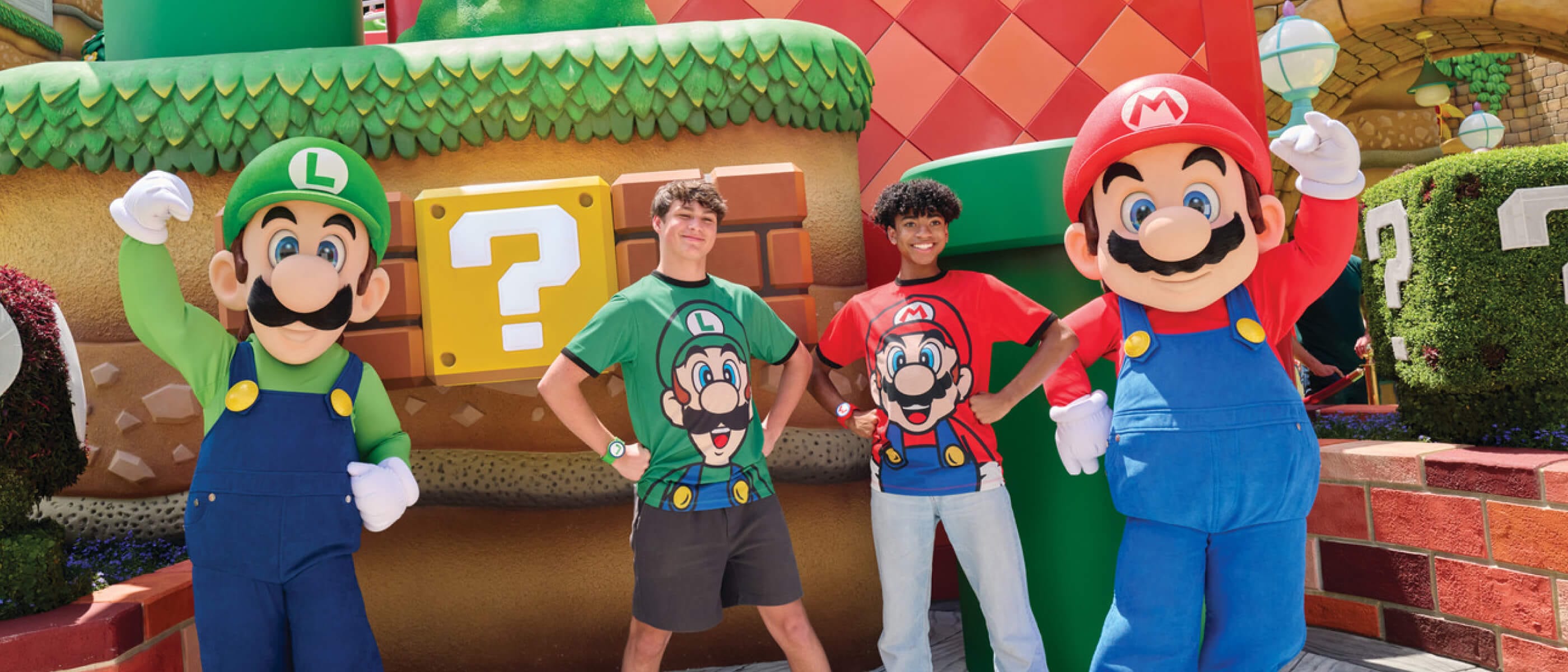 Dos amigos con playeras de Mario y Luigi posan con orgullo con Mario y Luigi en el colorido SUPER NINTENDO WORLD™.