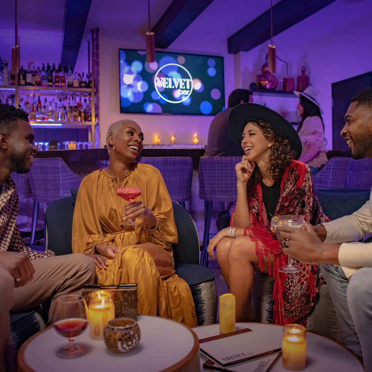 Cuatro amigos disfrutan de una variedad de cocteles rodeados de velas encendidas y el característico resplandor morado del Velvet Bar.