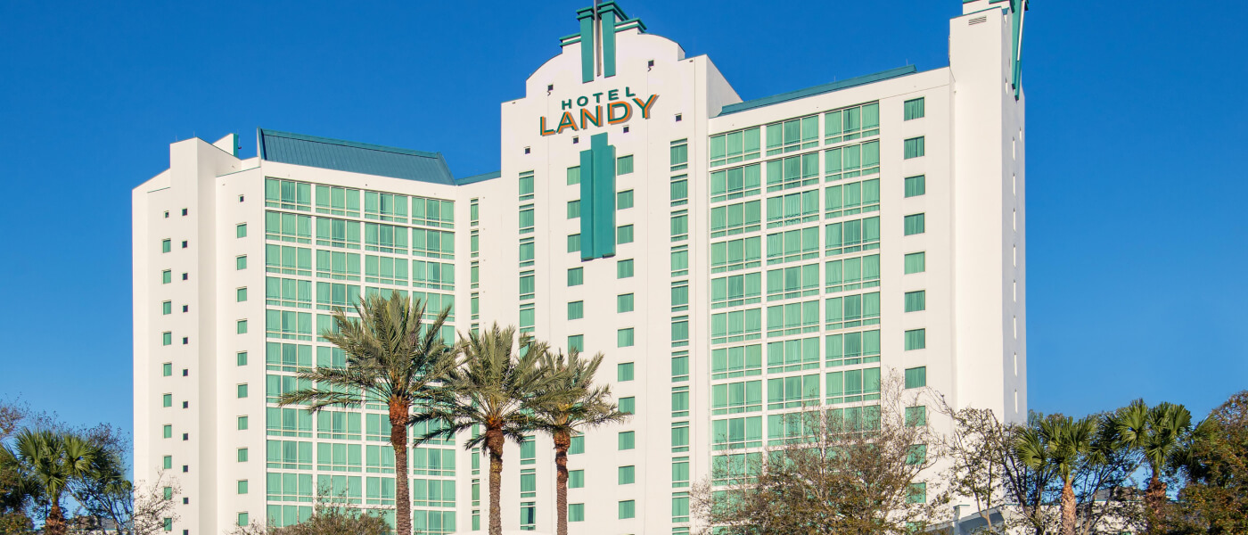 Large, light-colored building with green-tinted windows and a “Hotel Landy” sign at the top amid palm trees and a blue sky.