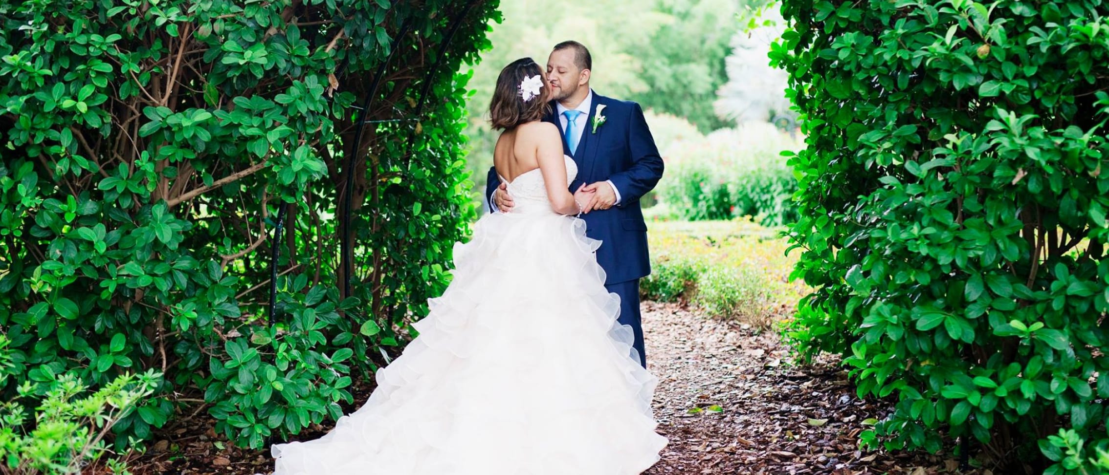 Una pareja de novios se da un beso frente a un arco con vegetación.