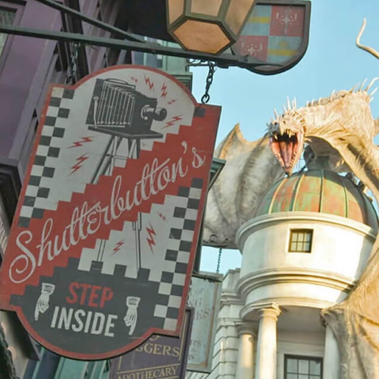 El letrero a la entrada de Shutterbutton’s™, el estudio de fotografía en The Wizarding World of Harry Potter™ - Diagon Alley™, y en el fondo se aprecia un dragón sobre el banco Gringotts™.