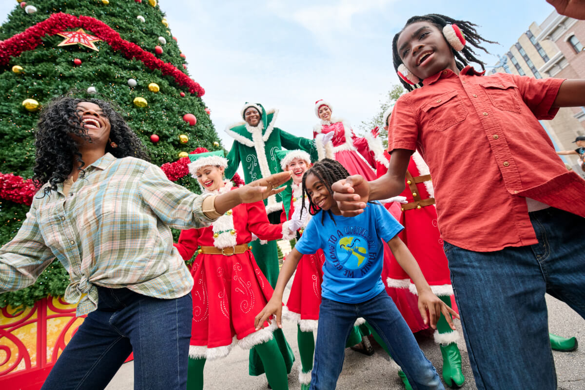 Una mamá y sus hijos bailan con unos elfos vestidos de rojo y verde junto a un gran árbol de Navidad en Universal Studios Florida.